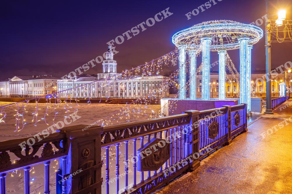 На фотографии изображен Дворцовый мост, украшенный яркими гирляндами, с видом на Стрелку Васильевского острова. На заднем плане виднеется здание Кунсткамеры, освещенное мягким светом. Замерзшая поверхность Невы подчеркивает зимнюю атмосферу, а украшенные колонны на мосту добавляют праздничное настроение. Это один из самых красивых видов Санкт-Петербурга в зимний сезон.